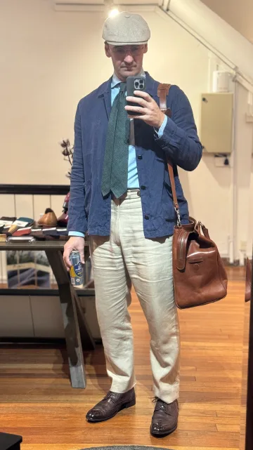 John Philip Green in a navy blazer, striped tie, cream trousers and flat cap, carrying a brown leather bag.