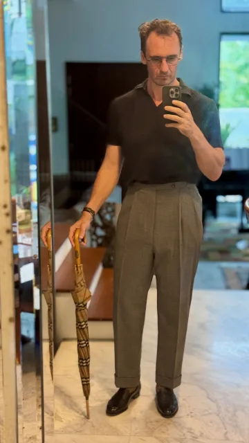 John Philip Green in a dark short-sleeved top and grey pleated trousers, holding an umbrella.