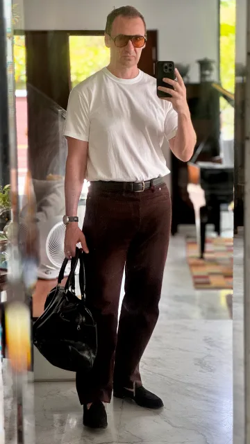 John Philip Green in a white T-shirt and brown trousers, carrying a dark leather bag.